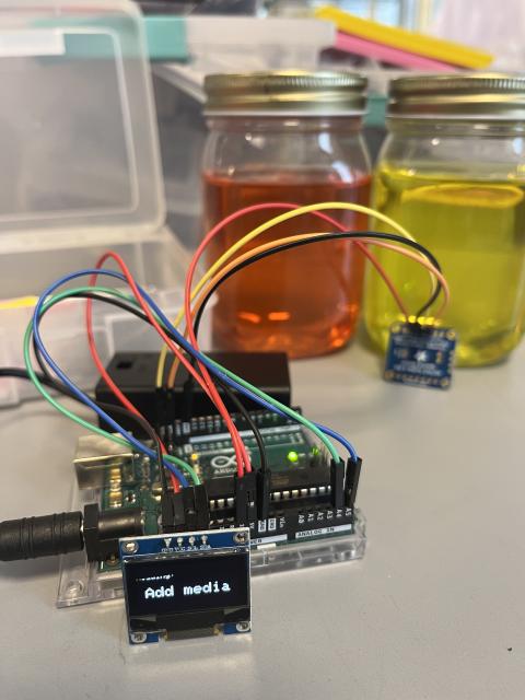 A circuit board connected to a sensor and OLED screen, reading and displaying the color of liquid in a jar.